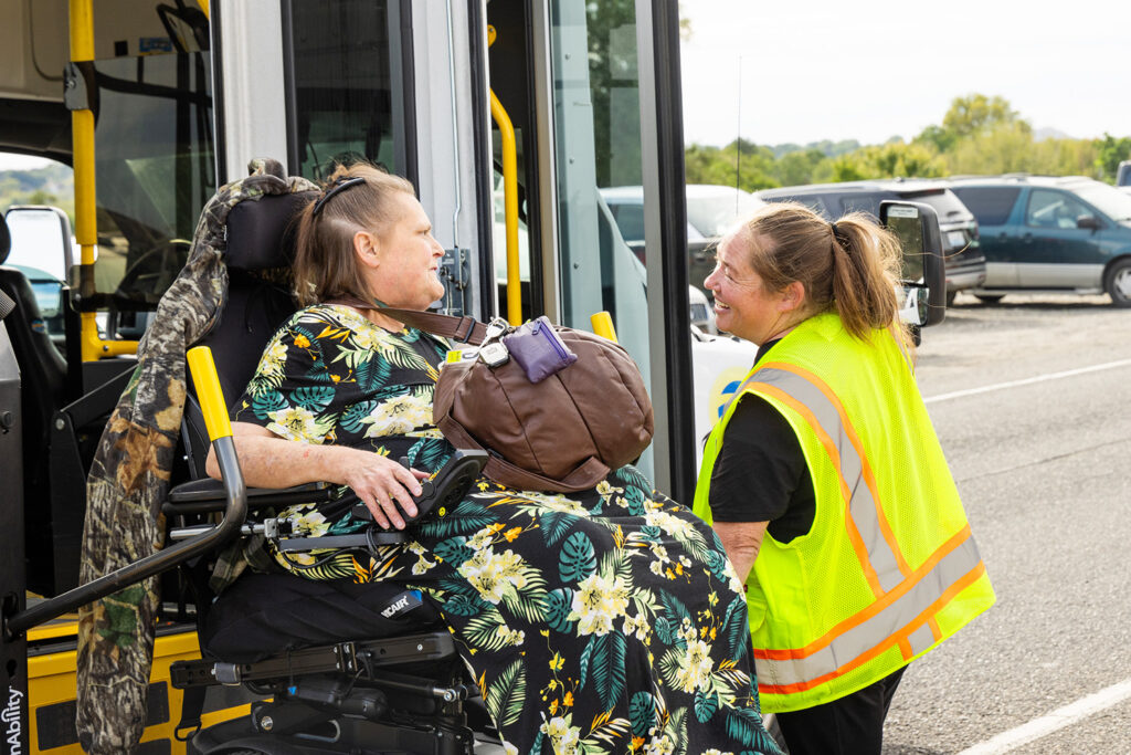A Dial-A-Ride driver smiles and assists a rider in a power wheelchair exit a Dial-A-Ride vehicle using the wheelchair lift.