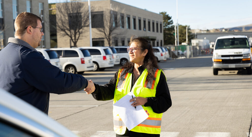 A BFT Rideshare employee smiles and shakes the hand of a Rideshare customer. They stand in the BFT lot and Rideshare vehicles can be seen in the background.